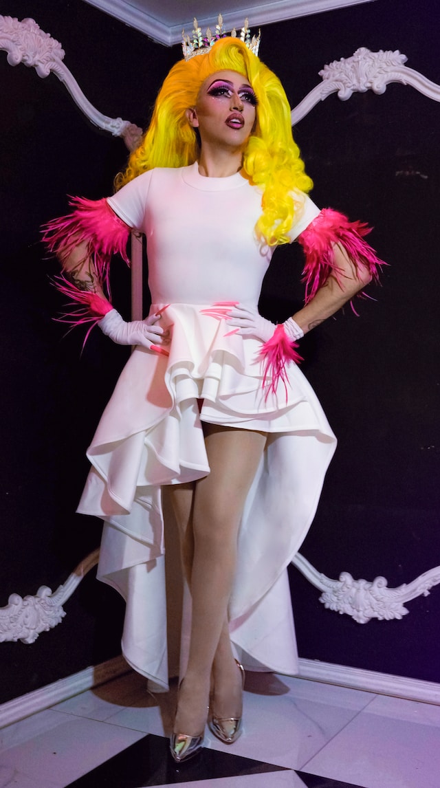 A drag queen wearing a pink dress with dark pink tassels and a bright yellow wig with a crown.