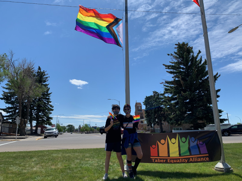 Two youth standing under the Progressive Prideflag in Taber - June 5, 2021.