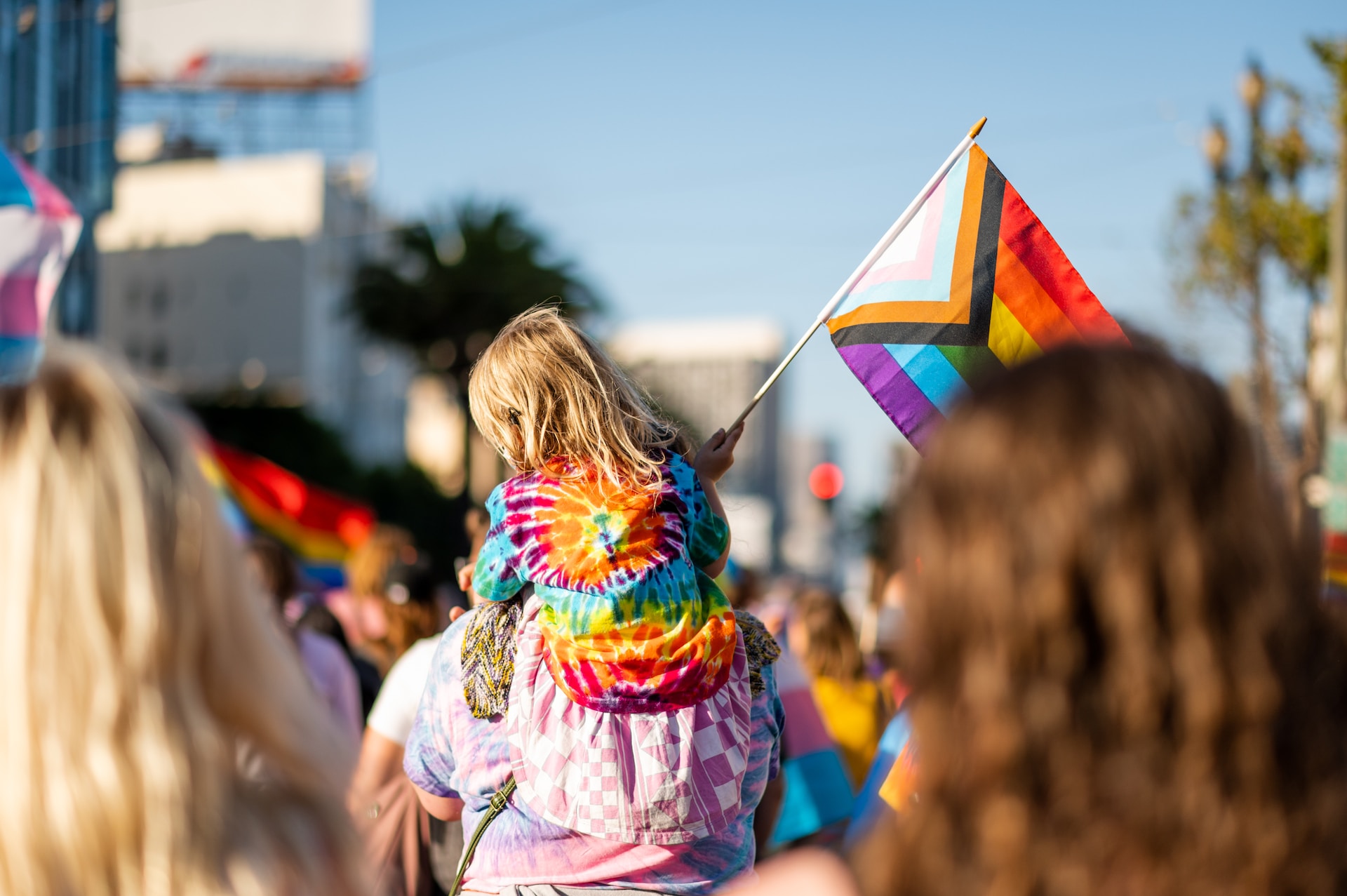 People in a 2SLGBTQIA+ march with a small child in a rainbow tie-died shirt riding on the back of an adult holding a progressive pride flag.