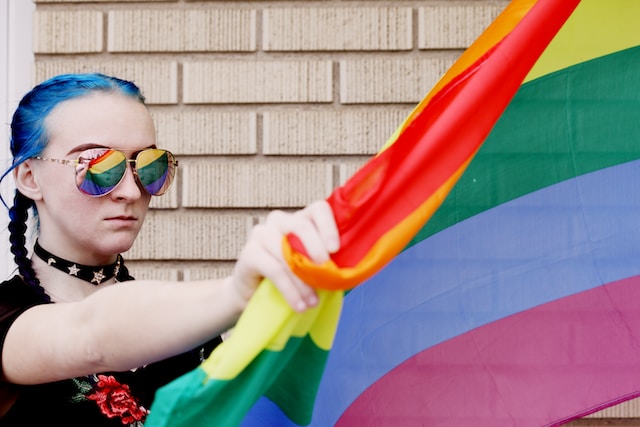 A photo of a youth holding a pride flag showing a reflection of the flag in their sunglasses.
